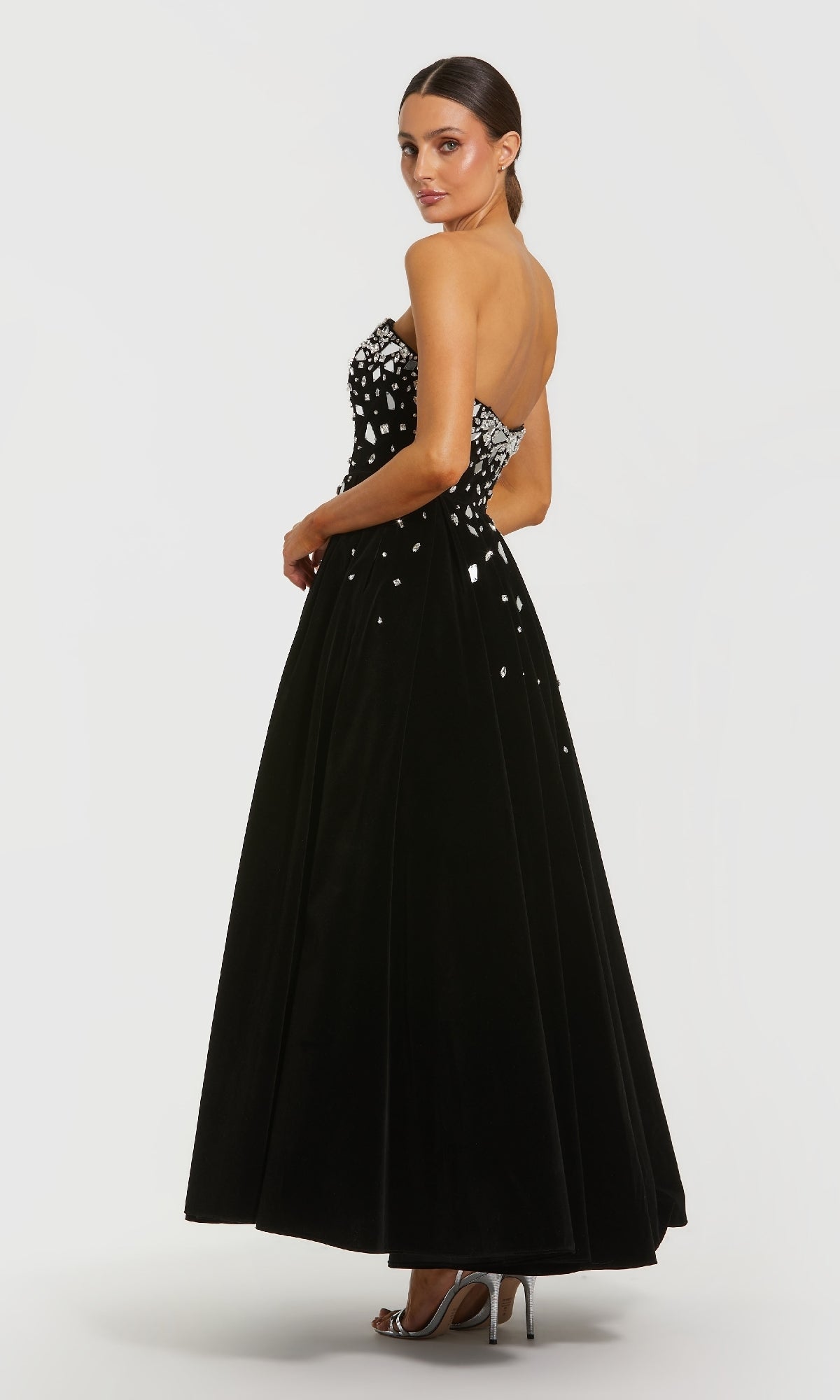 A woman in the Mac Duggal 56197 long formal dress—a strapless black velvet gown with a rhinestone bodice—pairs it with silver heels and stands against a plain white background.